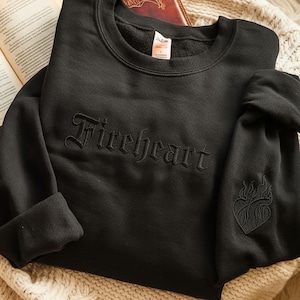May include: A black crewneck sweatshirt featuring the word Fireheart embroidered in a matching black gothic font across the chest. The left sleeve displays a small, tonal embroidered heart design with flame-like details on the lower arm.