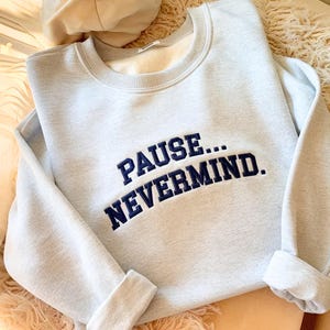 May include: A light gray crewneck sweatshirt featuring the dark blue embroidered text PAUSE... NEVERMIND. The garment has long sleeves with ribbed cuffs, a ribbed neckline, and a soft interior lining, presenting a clean and simple design.