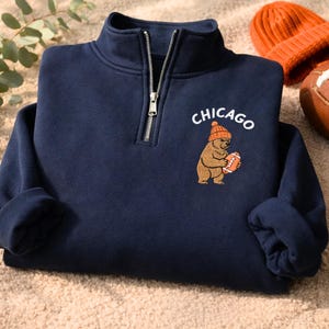 May include: Navy blue quarter-zip sweatshirt with a "Chicago" graphic featuring a bear wearing an orange beanie and holding a football. An orange beanie and a football are next to the sweatshirt.