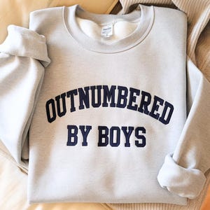 May include: A light gray sweatshirt with the text "OUTNUMBERED BY BOYS" in navy blue lettering. The sweatshirt has a crew neck and long sleeves, with the sleeves folded up. The garment is made of a soft material.