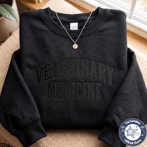 May include: A black crewneck sweatshirt featuring the words VETERINARY MEDICINE embroidered in a matching black thread across the chest in a collegiate font. A thin silver chain necklace with a small round pendant rests at the neckline.