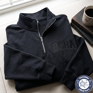 May include: A black quarter-zip sweatshirt featuring a high collar and a silver-toned metal zip. The front of the garment displays the letters BCBA embroidered in a matching black thread, with ribbed cuffs and a soft, fleece-lined interior texture.