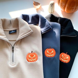 May include: Close-up of three quarter-zip sweatshirts in beige, blue, and black. Each sweatshirt features an embroidered orange jack-o'-lantern on the chest. The sweatshirts have a high collar and a silver zipper. The brand name is visible on the beige sweatshirt.