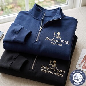 May include: A navy blue and a black quarter-zip sweatshirt, each featuring a gold embroidered skeleton graphic and personalized text. The navy sweatshirt reads Madison RT(R) Rad Tech, while the black sweatshirt reads Shelby RT(R) (ARRT) Diagnostic Imaging.