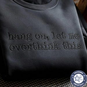 May include: A dark charcoal gray crewneck sweatshirt featuring the embroidered text "hang on, let me overthink this" in a matching dark thread color across the chest, creating a subtle, tonal appearance on the fabric surface.