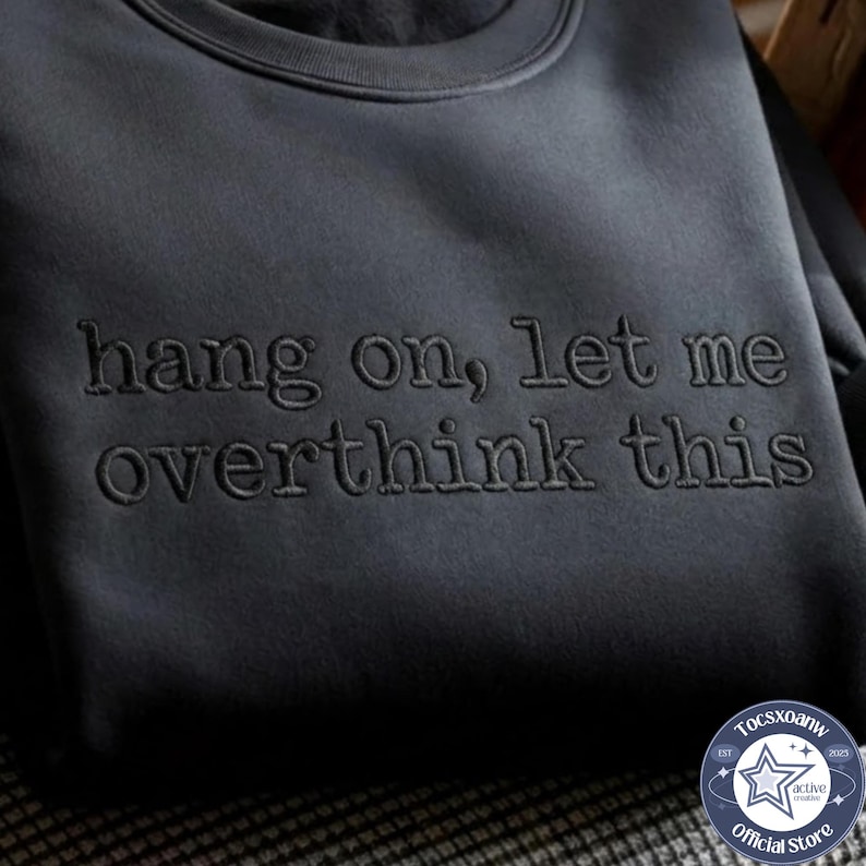 May include: A dark charcoal gray crewneck sweatshirt featuring the embroidered text "hang on, let me overthink this" in a matching dark thread color across the chest, creating a subtle, tonal appearance on the fabric surface.