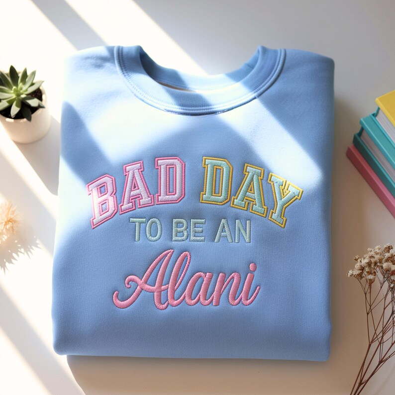 May include: A light blue sweatshirt with the embroidered text "BAD DAY TO BE AN Alani" in pink, yellow, and green. The sweatshirt is folded neatly on a white surface, with a small succulent plant and books in the background.
