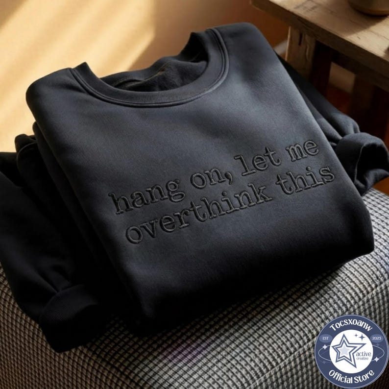 May include: A black crewneck sweatshirt featuring the embroidered text "hang on, let me overthink this" in a matching black thread. The garment has long sleeves, a ribbed collar, and a soft texture, showcasing a monochromatic design throughout.