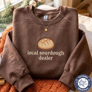 Peut inclure: Un sweat-shirt à col rond marron foncé avec une broderie représentant une miche de pain au levain ronde au-dessus du texte "local sourdough dealer" en lettres beige clair. Un collier à chaîne doré repose sur l'encolure du vêtement.