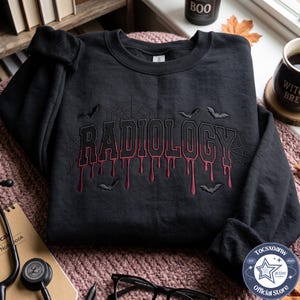 Puede incluir: Una sudadera negra de cuello redondo con un diseño bordado que muestra la palabra RADIOLOGY en una fuente de color rojo oscuro con efecto de goteo. El bordado incluye telarañas negras y pequeñas siluetas de murciélagos alrededor.