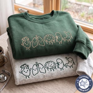 Puede incluir: Una sudadera verde oscuro y una sudadera gris jaspeado, cada una con un diseño bordado de líneas de cinco órganos humanos sonrientes: un corazón, pulmones, cerebro, riñón y estómago. Las sudaderas están apiladas una sobre la otra.
