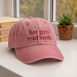 May include: A dusty rose baseball cap with the embroidered text "hot girls read books" in a darker red. The cap has a curved brim and a button on top. The hat is sitting on a white surface.