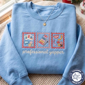 Peut inclure: Un sweat-shirt bleu clair à col rond orné de trois motifs de timbres brodés avec des icônes colorées incluant des photos, un xylophone, un téléphone et des pièces de puzzle, avec le texte professional yapper brodé en écriture cursive blanche.