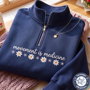 May include: A navy blue quarter-zip sweatshirt featuring a white embroidered daisy pattern and the text "movement is medicine" across the chest. The garment has a ribbed stand-up collar, ribbed cuffs, and a silver-toned metal zipper pull.