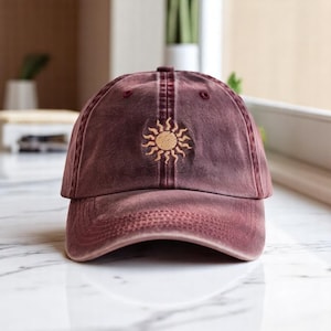 May include: A maroon baseball cap with a distressed look and a sun design embroidered on the front.
