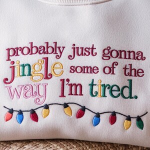 May include: Cream-colored sweatshirt with embroidered text reading "probably just gonna jingle some of the way I'm tired." The text is in various colors. A string of colorful Christmas lights is embroidered below the text.