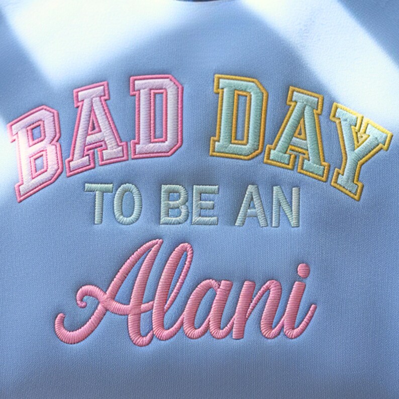 May include: Light blue sweatshirt with embroidered text. The text reads "BAD DAY TO BE AN Alani" in pink, white, and yellow lettering. The sweatshirt is a solid light blue color.