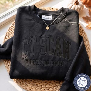 May include: A black crewneck sweatshirt featuring the text CT SCAN TECHNOLOGIST embossed in a collegiate font across the chest. The garment includes a white interior neck label reading ORIGINAL SWEATSHIRT CO. and a thin gold chain necklace.
