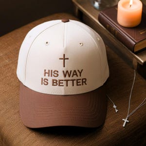 May include: A baseball cap with a white crown and a brown bill. The cap features the text "HIS WAY IS BETTER" embroidered in brown, along with a cross symbol. The cap is sitting on a brown surface.