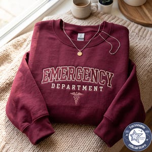 May include: A maroon crewneck sweatshirt featuring the white embroidered text EMERGENCY DEPARTMENT in an arched font, with a caduceus symbol below. A delicate gold-tone chain necklace with a small round pendant rests on the neckline.