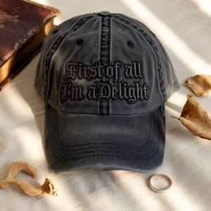 May include: A charcoal gray baseball cap with the embroidered text "First of all I'm a Delight". The hat has a curved brim and a distressed, vintage look. A gold ring and dried leaves are also visible.