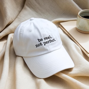 Könnte beinhalten: Weiße Baseballkappe mit dem gestickten Text "be real, not perfect." Die Kappe ist aus einem weichen Stoff gefertigt und hat einen gebogenen Schirm. Eine Keramik-Tasse mit dunkler Flüssigkeit und ein Stapel Papier befinden sich im Hintergrund.