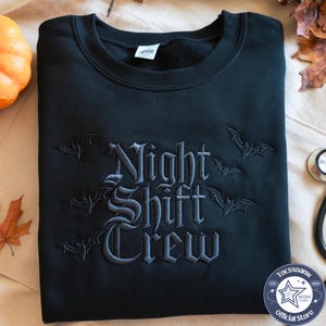 Puede incluir: Una sudadera negra de cuello redondo con texto bordado en gris oscuro que dice Night Shift Crew. El diseño está decorado con varias siluetas de murciélagos bordadas que rodean las letras en una fuente de estilo gótico en la parte frontal.
