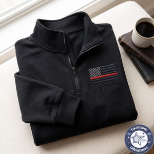 May include: A black quarter-zip sweatshirt featuring an embroidered American flag with a thin red line and the name Johnson stitched in black cursive script below the flag. The garment has a ribbed collar, long sleeves with ribbed cuffs, and a zipper.