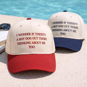 May include: Two baseball caps with the text "I WONDER IF THERE'S A HOT DOG OUT THERE THINKING ABOUT ME TOO". One cap has a red brim and the other has a navy blue brim. Both caps have a cream-colored crown.