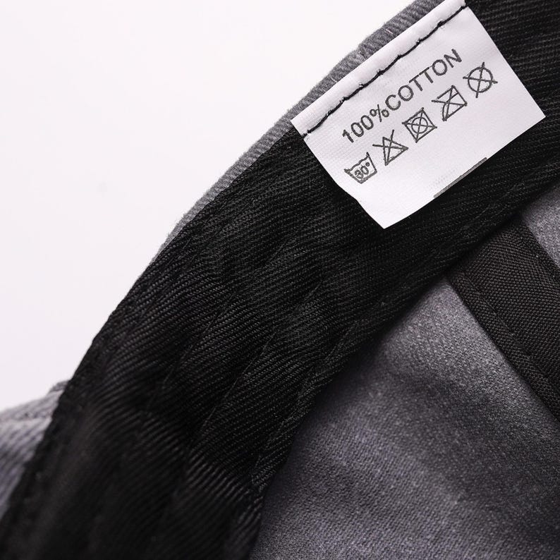 May include: Close-up of a gray baseball cap with a black interior and a white tag. The tag reads "100% COTTON" and includes washing instructions. The cap is made of a durable fabric.