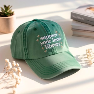 May include: A green baseball cap with the embroidered text "support your local library" in white. The hat has a curved brim and a fabric strap closure. The cap is made of a soft, durable material.