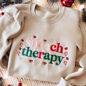 Peut inclure: Sweat-shirt beige avec l'inscription brodée "Speech therapy" en rose, vert et rouge, ornée de cœurs, flocons de neige et cannes en sucre. Un vêtement de fête et confortable.