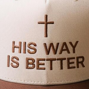 May include: A baseball cap with a brown brim and a cream-colored crown. The cap features a brown cross and the embroidered text "HIS WAY IS BETTER" in brown thread. The hat is a religious accessory.