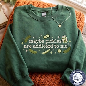 Puede incluir: Una sudadera de cuello redondo en color verde bosque con un diseño bordado de varios pepinillos y un frasco. El texto dice "maybe pickles are addicted to me", rodeado de pequeñas estrellas. Una fina cadena dorada descansa sobre el cuello.