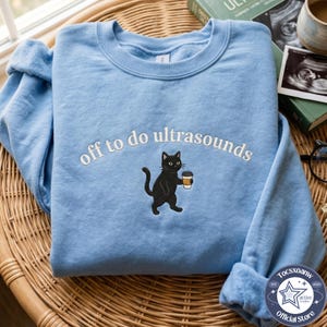 May include: A light blue crewneck sweatshirt featuring an embroidered black cat holding a coffee cup. The text "off to do ultrasounds" is embroidered in white above the cat. The design is centred on the chest of the long-sleeved garment.