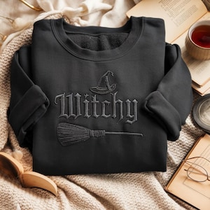 May include: Black sweatshirt with the word "Witchy" embroidered in a gothic font, along with a witch hat and broom design. The sweatshirt is folded on a cream-colored blanket, with a cup of tea and books in the background.