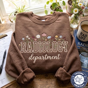 May include: A brown crewneck sweatshirt featuring embroidered colourful wildflowers above the text RADIOLOGY in a bold serif font and the word department in a cursive script. The design is centred on the chest of the long-sleeved garment.