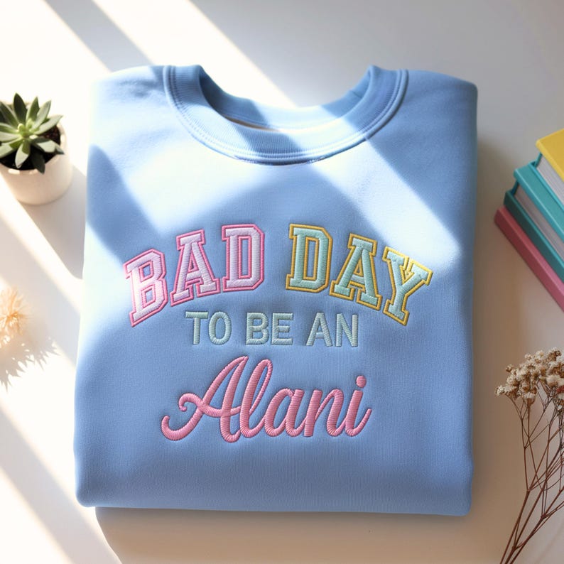 May include: A light blue sweatshirt with the embroidered text "BAD DAY TO BE AN Alani" in pink, yellow, and green. The sweatshirt is folded neatly on a white surface, with a small succulent plant and books in the background.