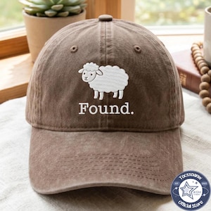 May include: A brown baseball cap featuring an embroidered white sheep graphic centered above the word Found. The cap has a structured crown, a curved brim, and a button on top. The design is stitched in white thread against the brown fabric.