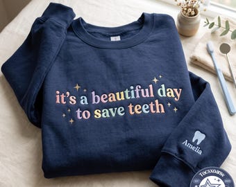 Può includere: Una felpa girocollo blu navy con il testo ricamato "it's a beautiful day to save teeth" in colori pastello e piccole stelle. La manica presenta un'icona di un dente bianco ricamata sopra il nome "Amelia" in caratteri bianchi.