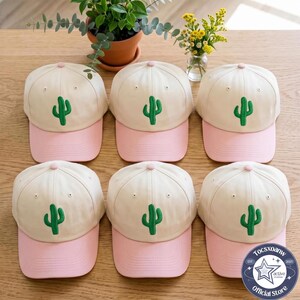 May include: Six baseball caps arranged in two rows, each featuring a cream-coloured crown with an embroidered green cactus and a solid light pink brim. Each cap has a small pink button at the top centre and eyelets for ventilation.