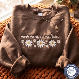 May include: A brown crewneck sweatshirt featuring the embroidered text "movement is medicine" in white cursive script above a row of white daisy flowers with yellow centers. A thin gold-toned chain necklace rests on the neckline of the garment.