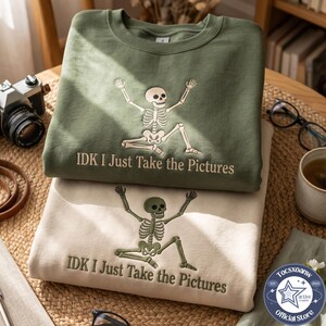 May include: Two crewneck sweatshirts, one olive green and one cream, each featuring an embroidered skeleton in a dynamic pose with arms raised and legs bent. Below the skeleton, the text reads IDK I Just Take the Pictures in matching thread.