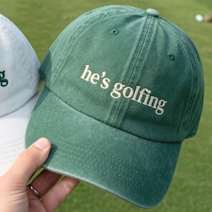 May include: A green baseball cap with the phrase "he's golfing" embroidered in white. The cap is made of a soft, textured fabric and has a curved brim. The cap is being held by a person.