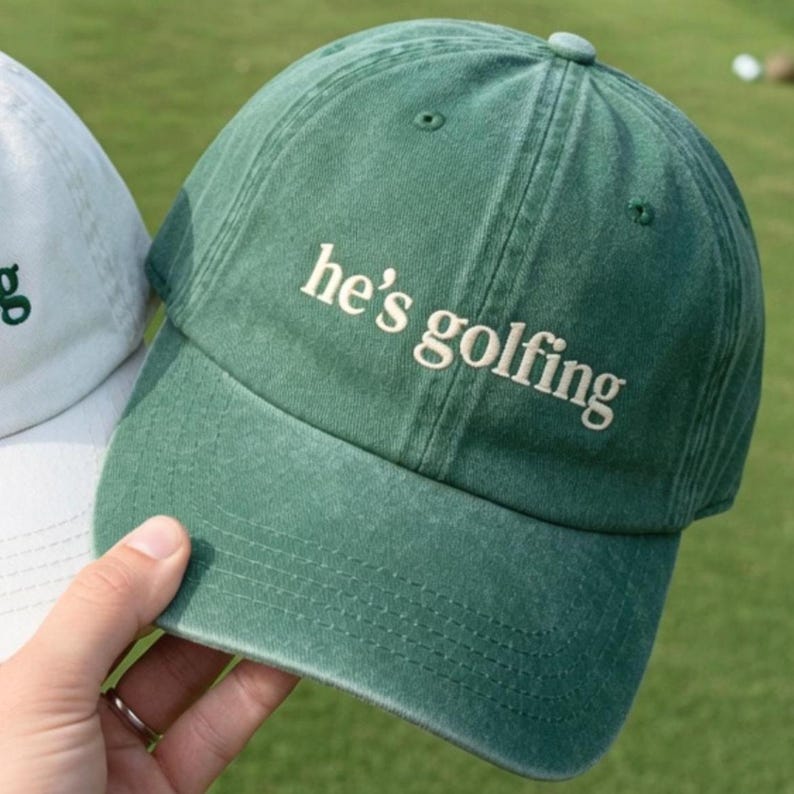 May include: A green baseball cap with the phrase "he's golfing" embroidered in white. The cap is made of a soft, textured fabric and has a curved brim. The cap is being held by a person.