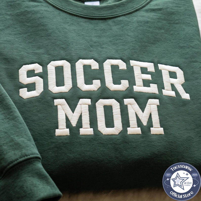 May include: A dark green crewneck sweatshirt featuring the text SOCCER MOM embroidered in large, white, block-style letters across the chest. The fabric has a visible texture, and the lettering is stitched with a raised, dense thread pattern.
