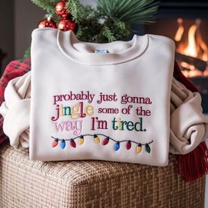 May include: Cream-colored sweatshirt with embroidered text reading "probably just gonna jingle some of the way I'm tired." The design includes colorful Christmas lights. The sweatshirt is folded on a woven basket.