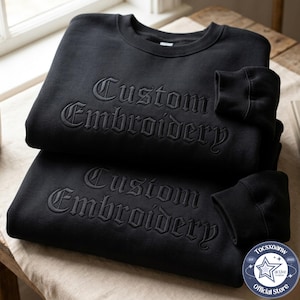 May include: Two black crewneck sweatshirts stacked on top of each other, each featuring the words Custom Embroidery stitched in a raised, tonal black gothic font across the chest. The garments have long sleeves with ribbed cuffs and a round neckline.