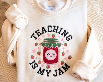 May include: Cream-colored sweatshirt with the embroidered phrase "TEACHING IS MY JAM." The design features a smiling jar of strawberry jam, surrounded by strawberries and hearts. The long-sleeved top is made of a soft material.
