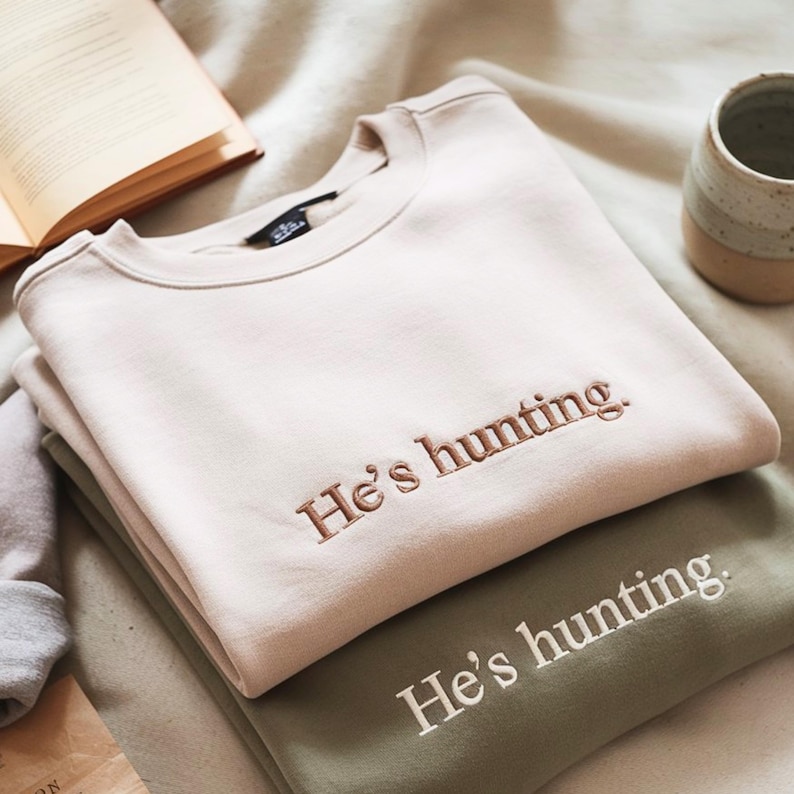 May include: Two sweatshirts, one beige and one olive green, are stacked. The beige sweatshirt has the embroidered text "He's hunting." in a brown thread. The olive green sweatshirt has the same text embroidered in white. The sweatshirts are on a soft, neutral-toned surface.